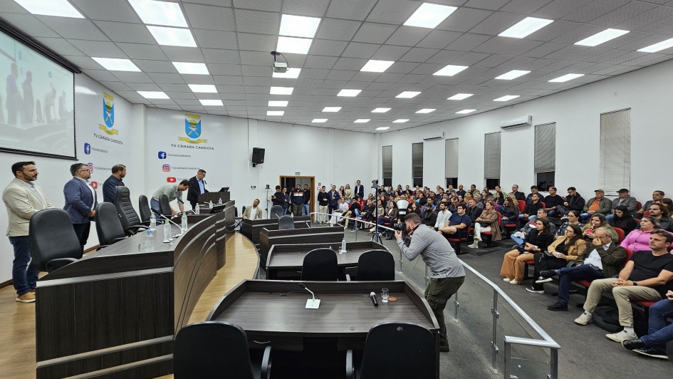 Foto ambiental de local fechado da audiência. À esquerda, uma mesa de apresentação com pessoas de pé junto a ela. À direita, o público assistindo sentado.