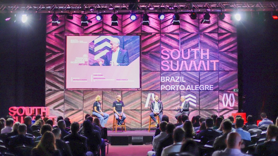 Foto da plateia de costas e de quatro pessoas sentadas sobre um palco com uma enorme tela atrás debatendo durante o o painel sobre Investimento em startups.
