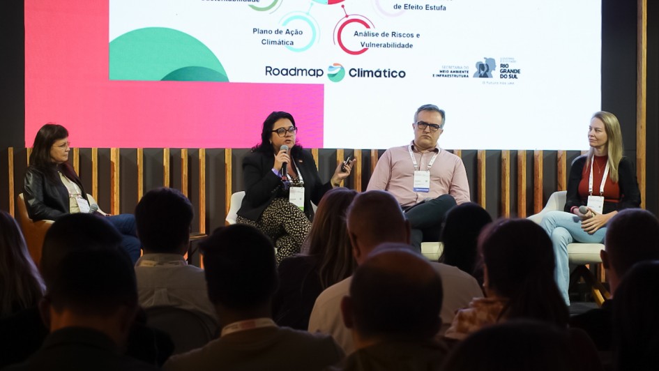 Foto do palco com os palestrantes sentados. Marjorie fala ao microfone. Na parte inferior da foto, é possível ver, pelas costas, as pessoas assistindo sentadas à apresentação.