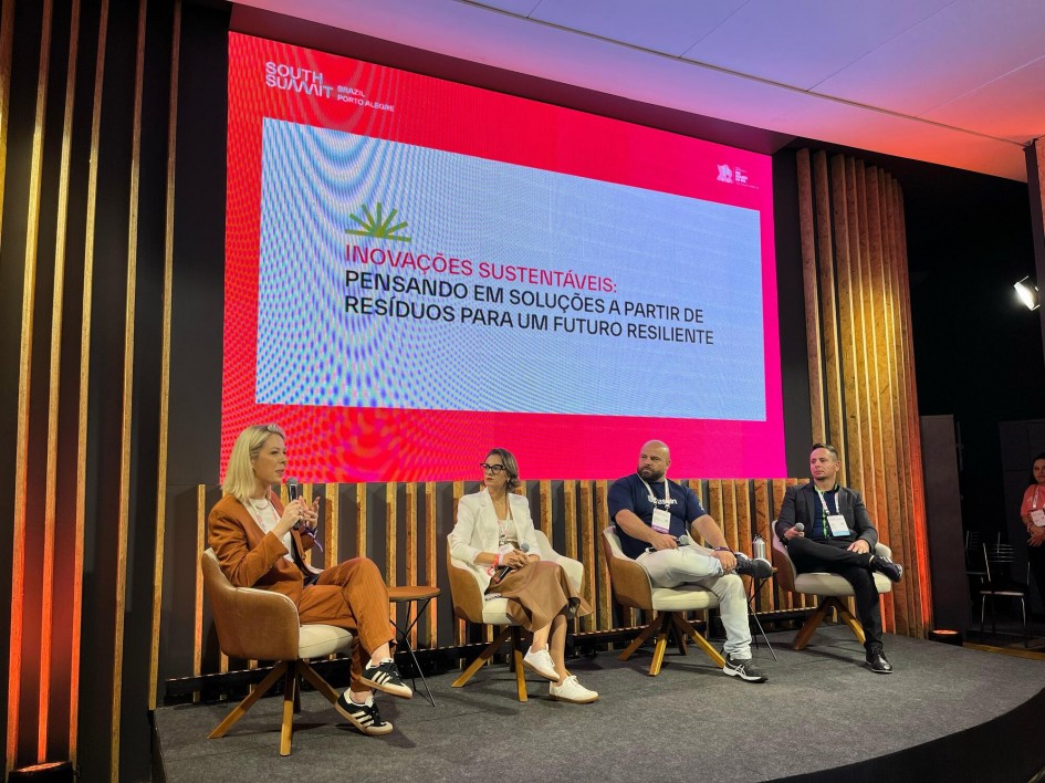 Foto meio lateral do palco com duas mulheres e dois homens sentados, um ao lado do outro e de frente pra plateia. Uma mulher segura o microfone. Atrás, está uma tela grande com o nome do painel sobre fundo branco.