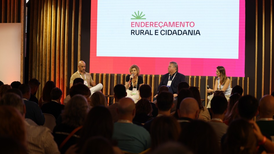 A imagem mostra quatro palestrantes (dois homens e duas mulheres), sentados em frente a um palco. Ao fundo há um painel branco e cor de rosa com a seguinte inscrição Endereçamento Rural e Cidadania. Há uma plateia presente.