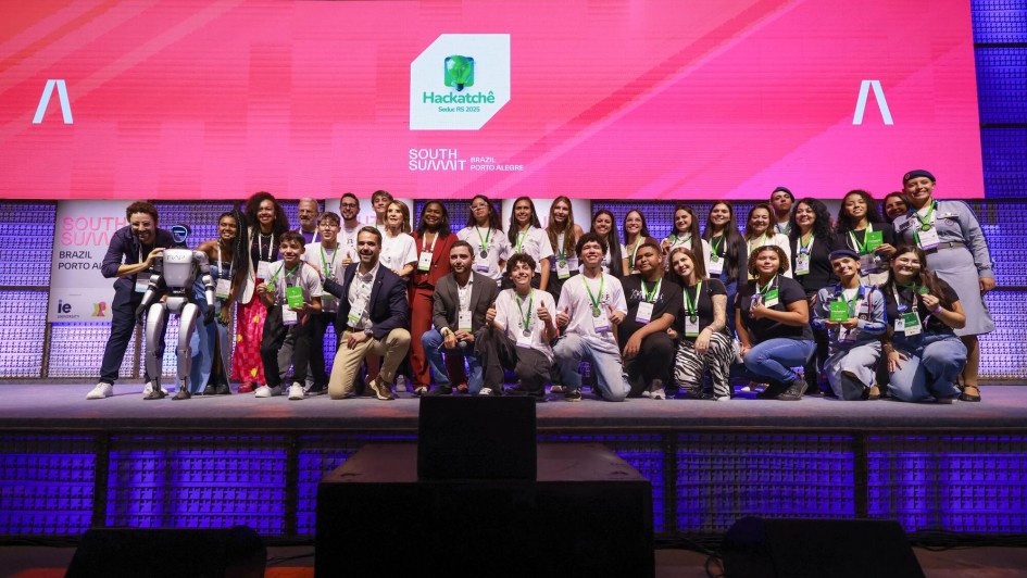 Um grande grupo de jovens e adultos posa no palco do evento "South Summit Brazil" em Porto Alegre. Ao centro, há um robô humanoide. Todos estão sorrindo, usando crachás e medalhas, indicando uma premiação ou reconhecimento. O telão exibe o logo do Hackatchê 2025 e o nome do evento.