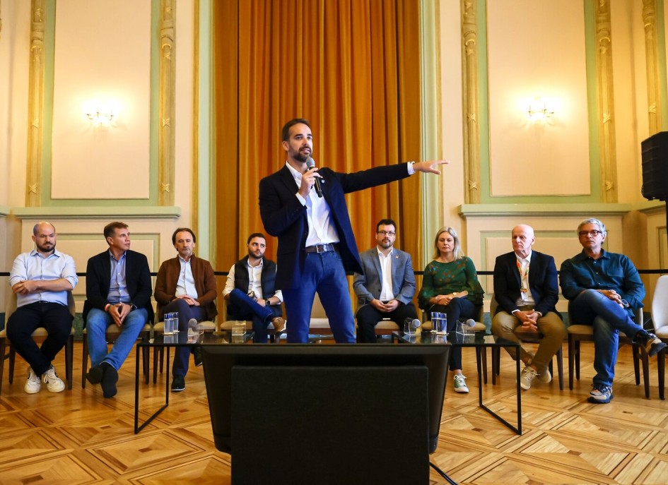 Leite em pé gesticulando e falando ao microfone. Atrás dele estão sentados oito secretários de estado, na reunião com prefeitos sobre o Bloco 2 de concessões.