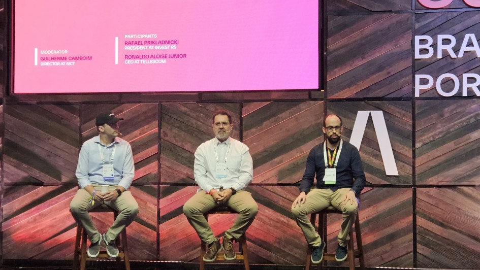 Foto em ambiente fechado. Há três homens sentados em banquetas em um palco, vestindo roupas sociais. Ao fundo, há um painel cor de rosa com a projeção dos nomes dos palestrantes.