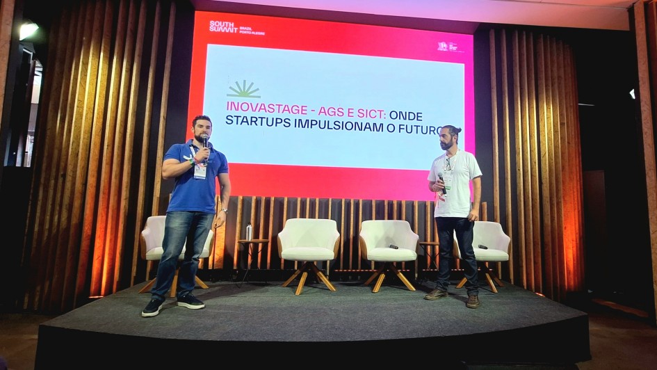 Foto em ambiente interno. Há dois palestrantes em pé em um palco. Ao fundo há um painel branco e cor de rosa, com a seguinte inscrição: "Inovastage - AGS e Sict: onde startups impulsionam o futuro". 
