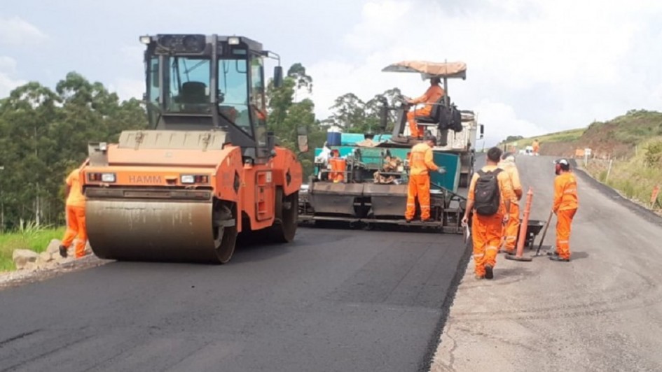 Há homens vestindo roupas laranjas andando no asfalto. Há uma máquina pavimentadora de asfalto. E uma pessoa está sentada numa cadeira em cima de uma caçamba.