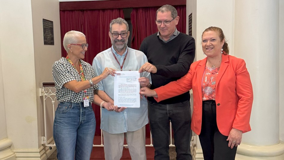 H&aacute; quatro pessoas e todas elas seguram um papel. Atr&aacute;s delas h&aacute; uma cortina bord&ocirc;. Tr&ecirc;s pessoas est&atilde;o de &oacute;culos. 