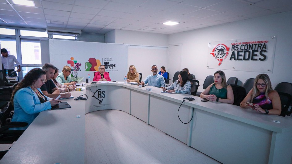 Mesa de reunião em U na cor branca, numa sala de piso e paredes também brancos. Há aproximadamente dez pessoas sentadas junto à mesa. Ao fundo, na parede, há uma placa em que está escrito RS contra Aedes.