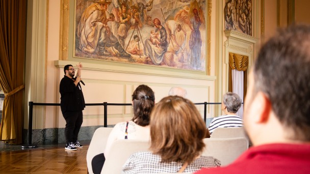 Um homem de barba e óculos, vestido de preto, segura um microfone e gesticula enquanto fala para uma plateia sentada. Ele está em pé, em frente a um grande painel pintado na parede, que retrata uma cena rural com várias pessoas em trajes antigos. O ambiente é elegante, com cortinas douradas, piso de madeira brilhante e detalhes clássicos na arquitetura. A plateia, composta por pessoas adultas e idosas, está atenta à apresentação.