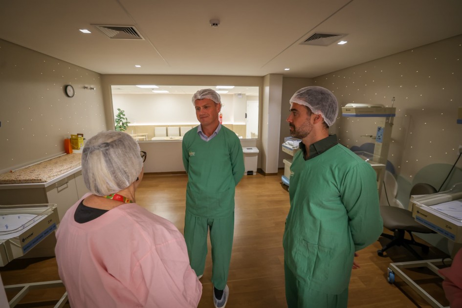 Três pessoas estão de pé em uma sala hospitalar similar à da primeira imagem. Dois homens, um deles o governador, usam roupas verdes de visita hospitalar e toucas protetoras, enquanto Arita está de costas usa jaleco rosa e também uma touca. Eles conversam em frente aos berços hospitalares da sala de obstetrícia.