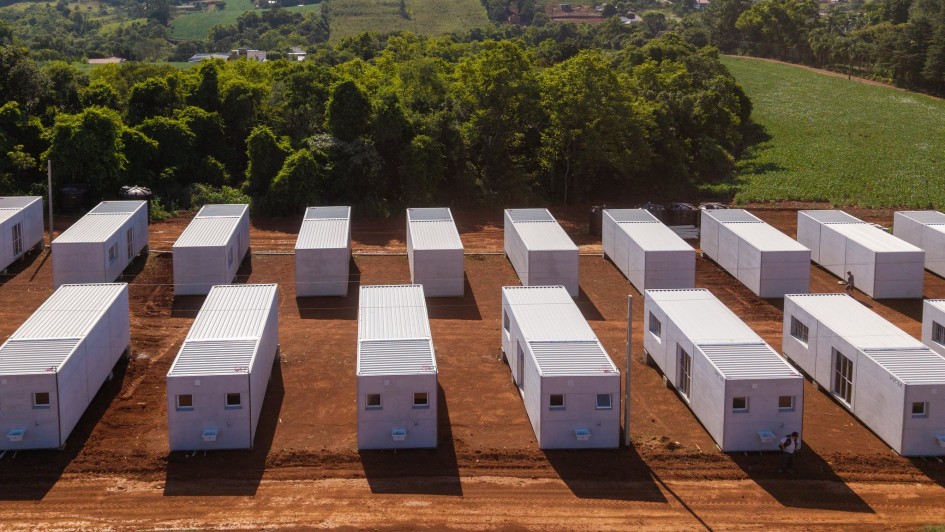 Há várias casas temporárias em um terreno de chão batido. No fundo, um morro com árvores e gramado.