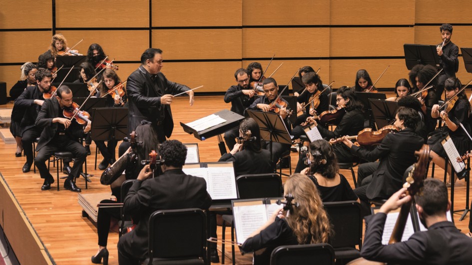 Há uma orquestra no palco. As pessoas estão sentadas com diversos instrumentos nas mãos sendo comandadas por um regente que está em pé.