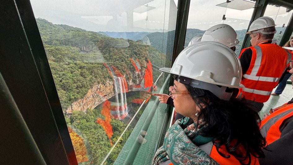 Há pessoas usando capacete e olhando para uma janela. A vista tem um morro com árvores. Elas usam colete de sinalização na cor laranja.