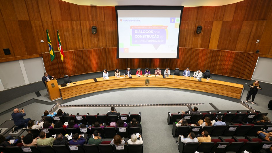 Há uma bancada com nove pessoas sentadas e uma pessoa de pé falando no microfone. Em frente, há diversas pessoas sentadas assistindo. Ao fundo, existe um telão de apresentação.