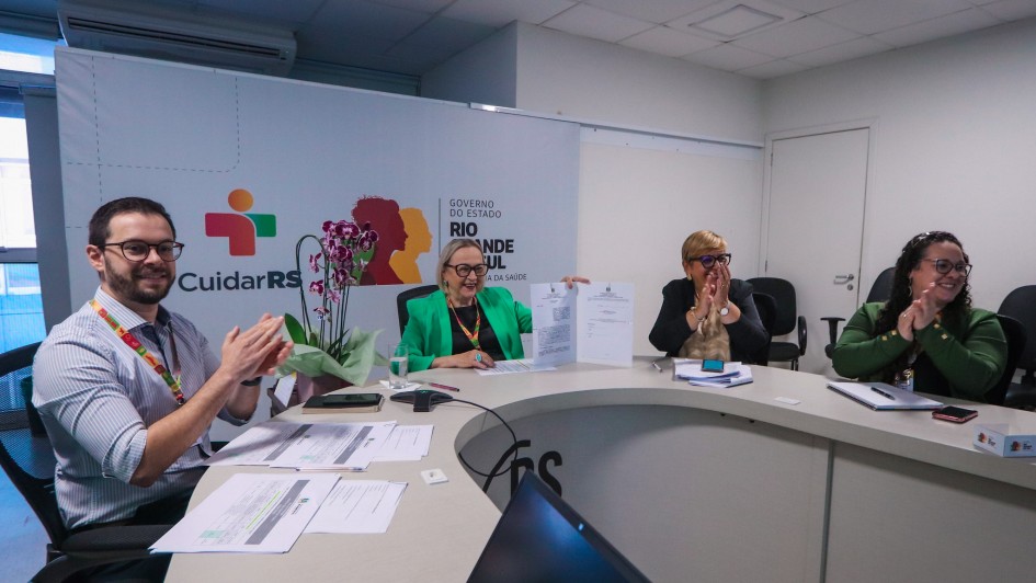 Há quatro pessoas sentadas numa bancada e sorrindo. Três estão batendo palmas e uma uma página aberta. É uma sala de cor clara. No fundo há um banner com a descrição e a logo do Estado do Rio Grand edo Sul.