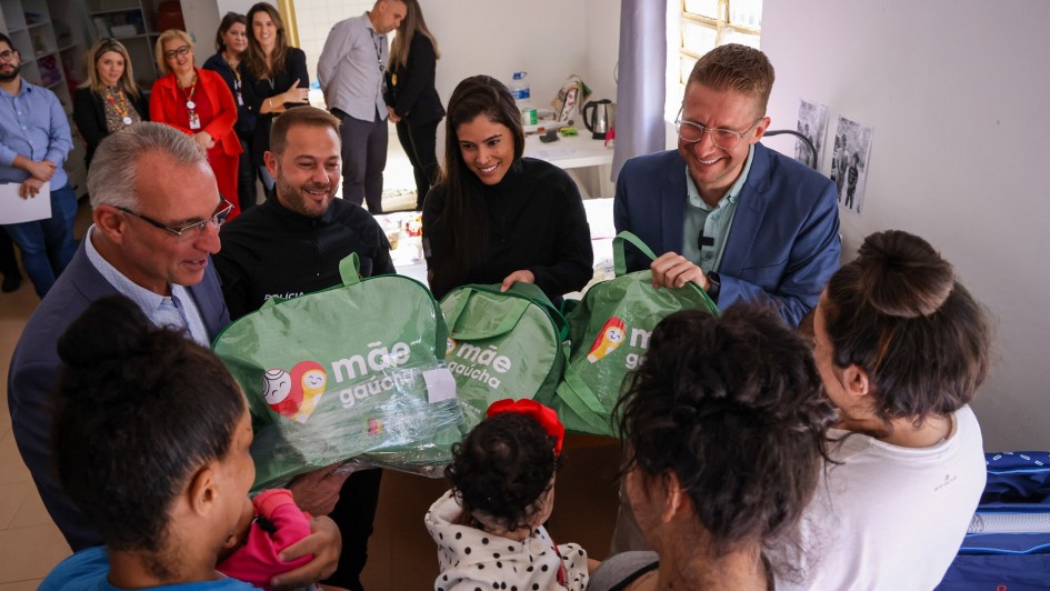 Na foto há varias pessoas de pé. Há quatro pessoas entregando três sacolas verdes para três mulheres que estão de costas. Uma delas segura um nenê.