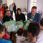 Na foto há varias pessoas de pé. Há quatro pessoas entregando três sacolas verdes para três mulheres que estão de costas. Uma delas segura um nenê.