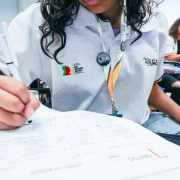 Há quatro alunos que aparecem na foto. Três estão ao fundo. A que está no primeiro plano aparece escrevendo com uma caneta em um papel.