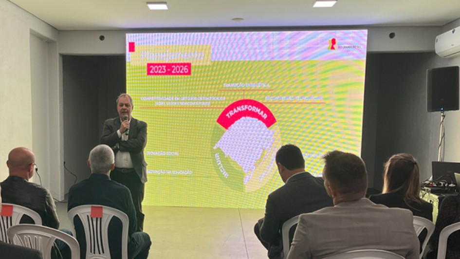 Há pessoas sentadas assistindo uma palestrante. No fundo, há um banner nas cores vermelha, verde e amarela com informações.