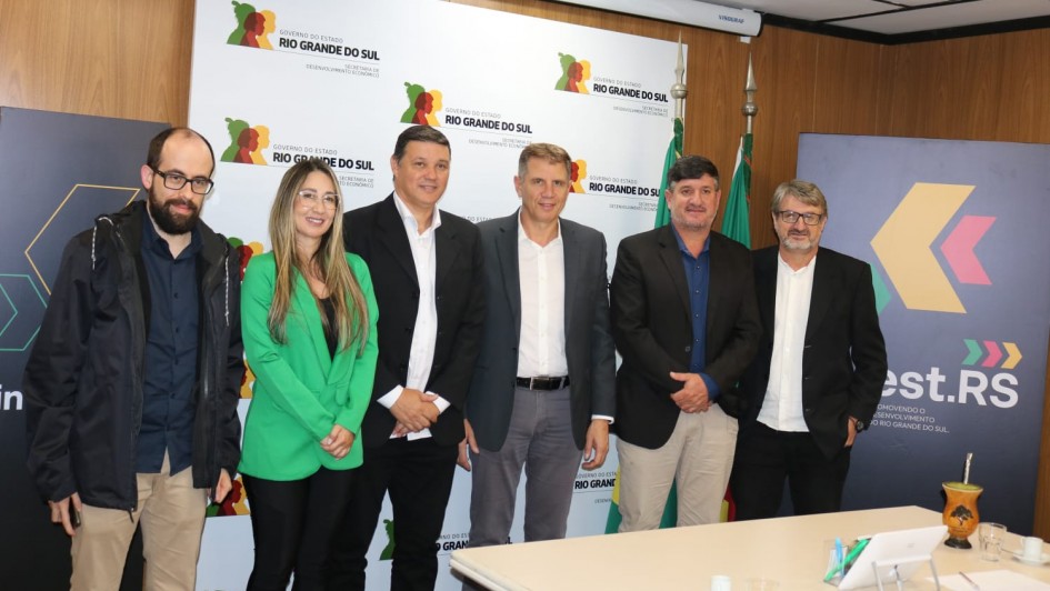 Foto posada de cinco pessoas numa sala. Elas estão de frente para a câmera, com um painel ao fundo do governo do RS.
