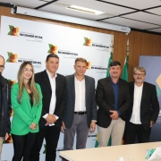Foto posada de cinco pessoas numa sala. Elas estão de frente para a câmera, com um painel ao fundo do governo do RS.