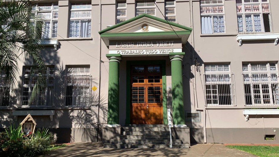 Há uma imagem de uma escola de cor cinza com várias janelas abertas. Há degraus até a porta de entrada .