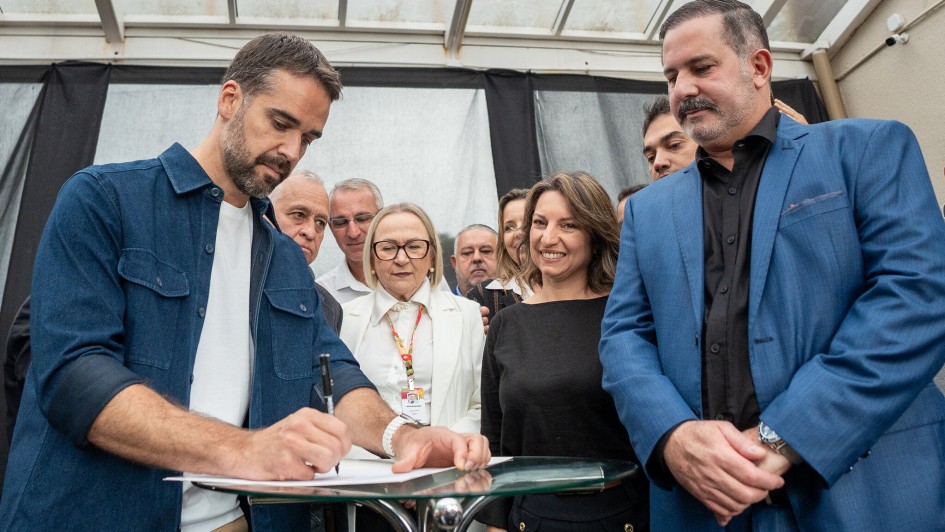 Leite assinando o anúncio de investimentos na saúde em Santiago sendo observado pelas pessoas que estão à sua volta.