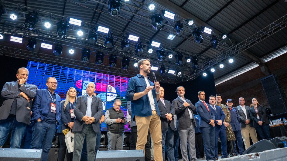 Leite sobre um palco, em pé, com um microfone, falando para plateia na abertura da Fecoarti 2025. Diversas pessoas estão junto com ele no palco.