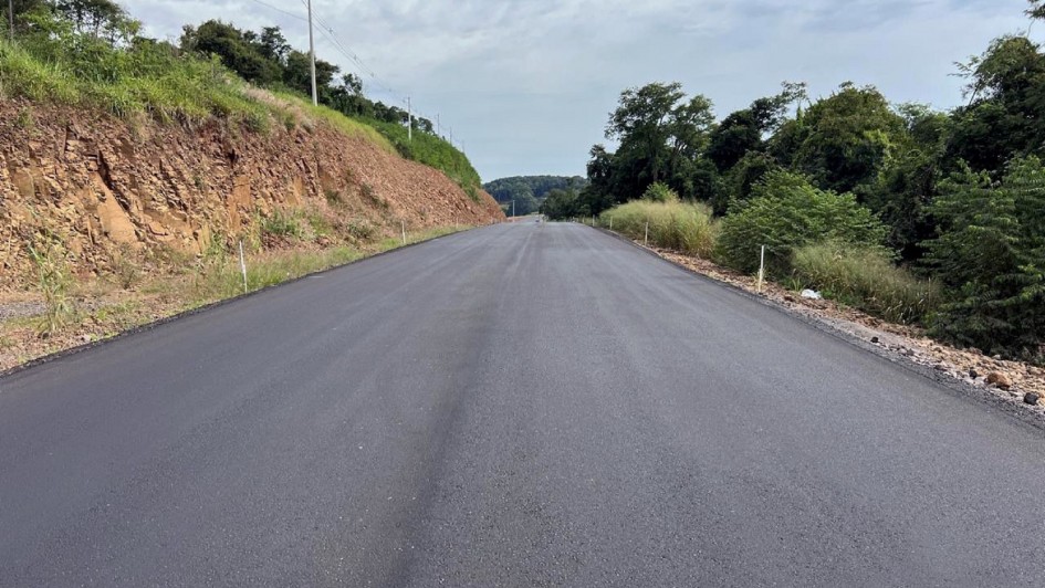 Na imagem há uma rua asfaltada. Do lado direito aparecem arbustos e do lado esquerdo um morro de média altura.
