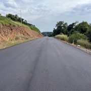 Na imagem há uma rua asfaltada. Do lado direito aparecem arbustos e do lado esquerdo um morro de média altura.