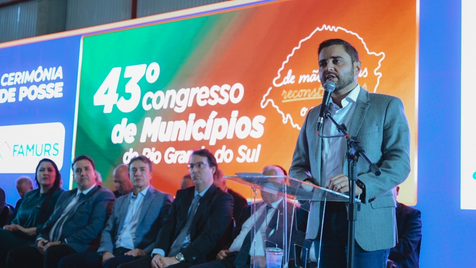 Há uma pessoa de pé falando ao microfone e cinco pessoas na fileira da frente sentadas. Ao fundo há um banner colorido.
