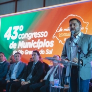 Há uma pessoa de pé falando ao microfone e cinco pessoas na fileira da frente sentadas. Ao fundo há um banner colorido.