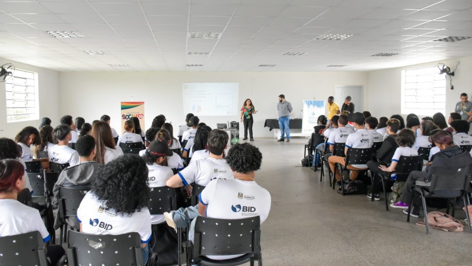 Em uma sala, há dezenas de alunos sentados de costas olhando para frente. Existem 5 pessoas de pé. Há duas janelas na sala, uma em cada parede lateral. Ao fundo ocorre uma apresentação.