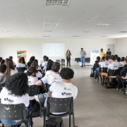 Em uma sala, há dezenas de alunos sentados de costas olhando para frente. Existem 5 pessoas de pé. Há duas janelas na sala, uma em cada parede lateral. Ao fundo ocorre uma apresentação.