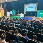 Em um auditório há dezenas de crianças sentadas assistindo uma apresentação. No palco há um palestrante e três banners. O fundo tem cortinas escuras e do lado esquerdo há uma parede marrom.