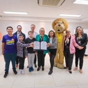 Foto posada da secretária segurando o documento assinado e exibindo para a câmera, ao lado de crianças e outros adultos. Há uma pessoa fantasiada de leão, um mascote da causa.