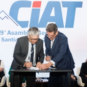 Dois homens assinando um papel que está sobre uma mesa sendo observados por duas mulheres sentada um pouco mais atrás. Trata-se da solenidade de assinatura de termo de cooperação entre a Sefaz e o Ciat no Chile 2025.