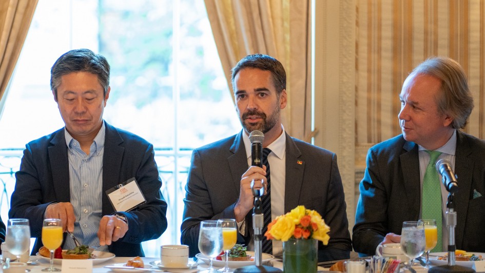 Na imagem aparecem três pessoas. Uma segura o microfone. Uma está com talheres nas mãos. E a outra pessoas está olhando para o lado. Todos estão de terno.