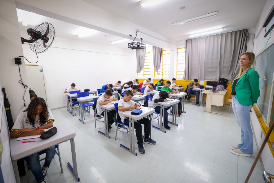 Várias crianças estão numa sala de aula sentadas e de frente para as classes. Há uma mesa grande ao fundo. Uma mulher de pé, em frente à turma. Há um ventilador de teto e um projetor na sala, além das cortinas cinzas. A cor das paredes da sala são brancas.