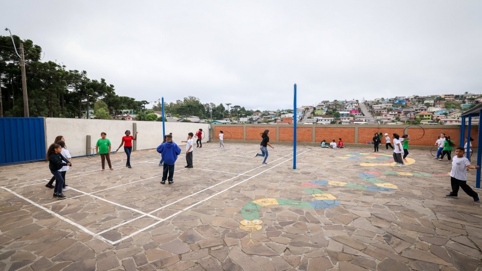 Há um pátio com dezenas de crianças brincando. O muro em volta é de tijolo à vista, parede branca e com um portão na cor azul. No chão há pinturas de desenhos coloridos e uma marcação de quadra de vôlei.