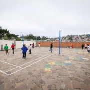 Há um pátio com dezenas de crianças brincando. O muro em volta é de tijolo à vista, parede branca e com um portão na cor azul. No chão há pinturas de desenhos coloridos e uma marcação de quadra de vôlei.