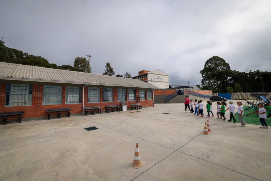 Há uma fila de crianças ao lado de um campo verde com com gangorra. Eles estão no pátio aberto de uma escola. Há cones de sinalização no pátio. E no fundo uma rampa de acesso a outra área da escola. Há uma pessoa de camisa vermelha que parece cuidar das crianças.