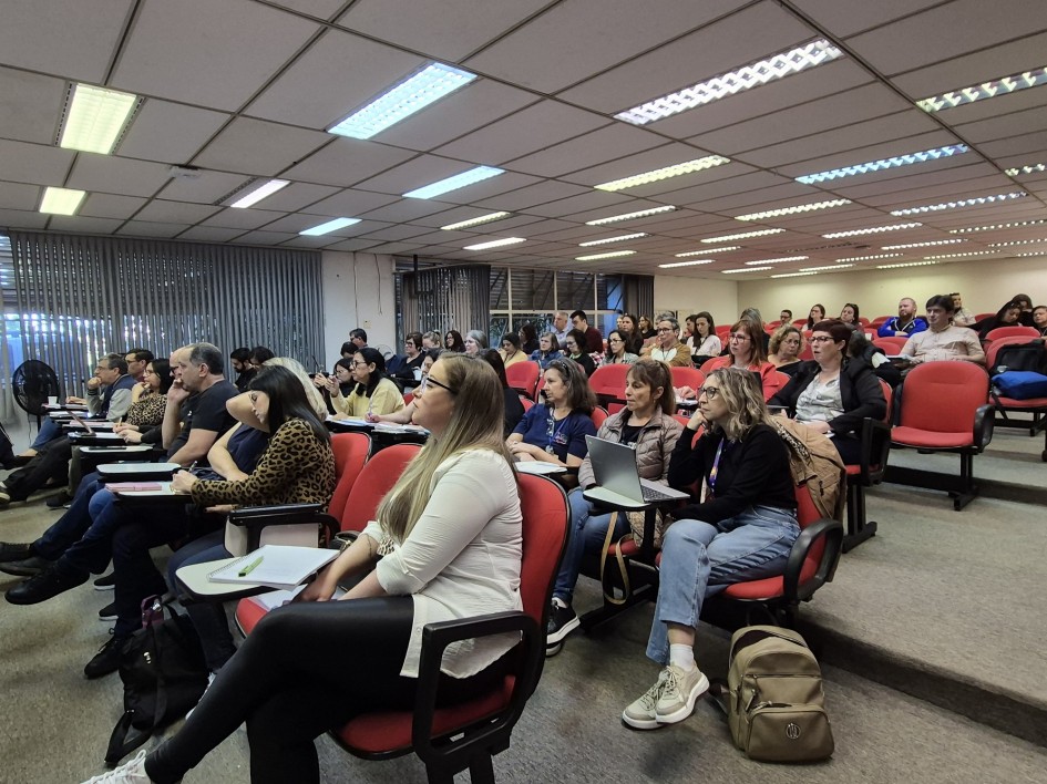 Há dezenas de pessoas sentadas em poltronas de cor vermelha em uma sala com diversos degraus. Há luzes no teto e janelas ao fundo.