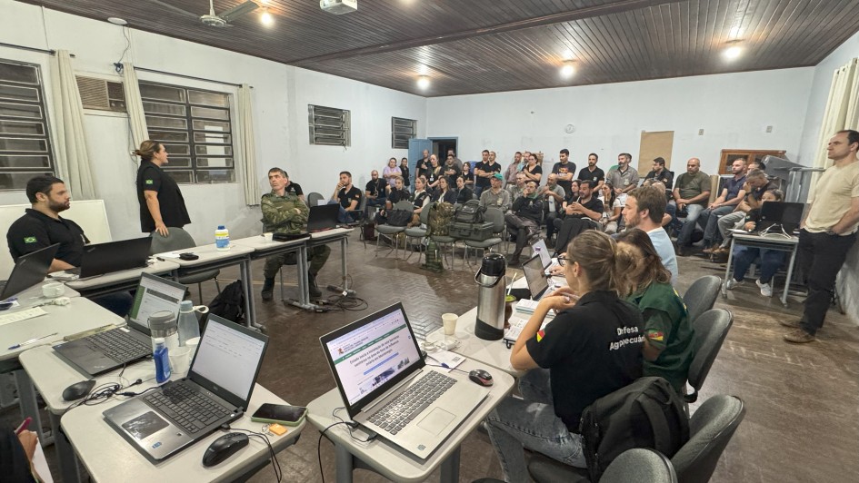 A foto mostra um grupo de pessoas reunidas em uma sala. Há mesas com laptops e pessoas sentadas, algumas olhando para a frente e outras trabalhando em seus computadores.