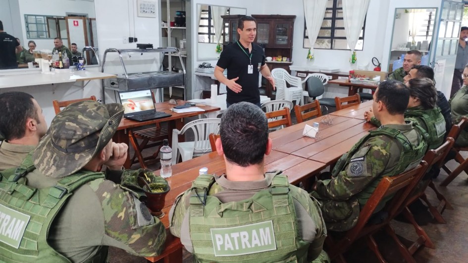 A foto mostra um homem de camisa preta gesticula em frente a um grupo de pessoas fardadas sentadas em mesas de madeira, algumas olhando para um laptop aberto.