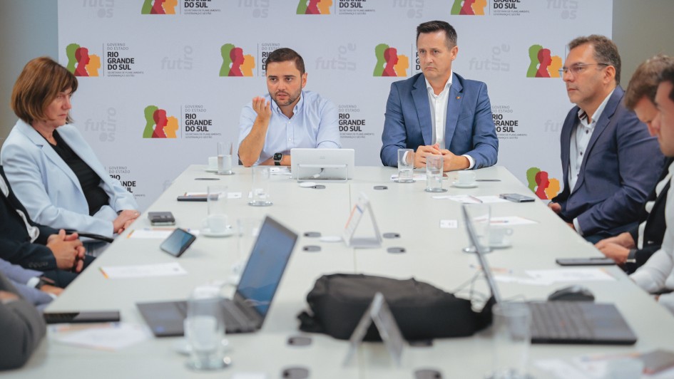 Há 8 pessoas sentadas. Em uma mesa retangular, há notebooks, celulares e copo de água. Três pessoas estão de terno. Ao fundo há um painel do Estado do Rio Grande do Sul.