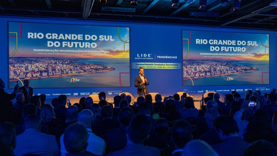 Em um palco de apresentação, há um homem de pé segurando um microfone. Dezenas de pessoas sentadas assistem ele. O palco tem a cor azul e há dois painéis grandes escrito Rio Grande do Sul do Futuro.