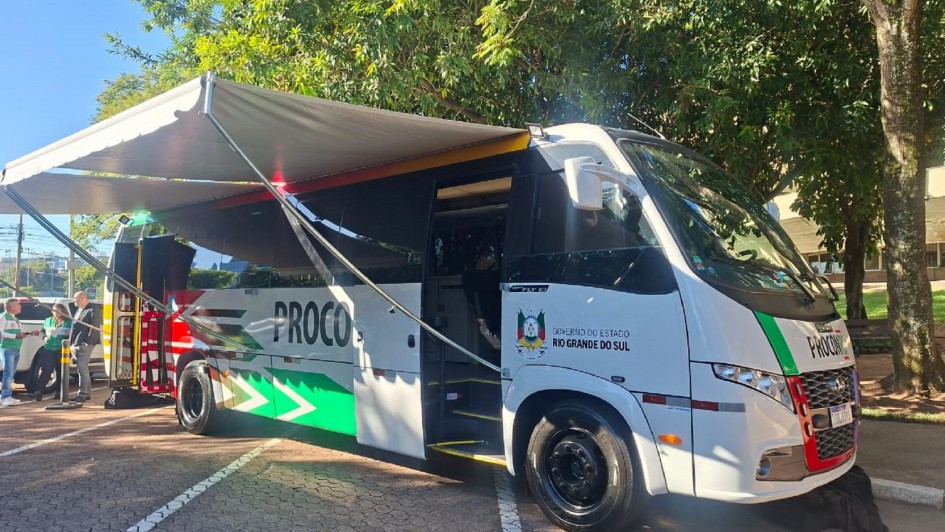 Há um micro-ônibus estacionado com a porta aberta e um toldo estendido para fazer sombra. Ao fundo, tem três pessoas de pé conversando. O ônibus é branco e possui a logo do estado do rio grande do sul.