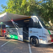 Há um micro-ônibus estacionado com a porta aberta e um toldo estendido para fazer sombra. Ao fundo, tem três pessoas de pé conversando. O ônibus é branco e possui a logo do estado do rio grande do sul.