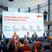 Há seis pessoas no palco. Cinco estão sentadas e uma de pé. Uma mulher no centro do palco está com o microfone na mão. Atrás deles há um painel. 